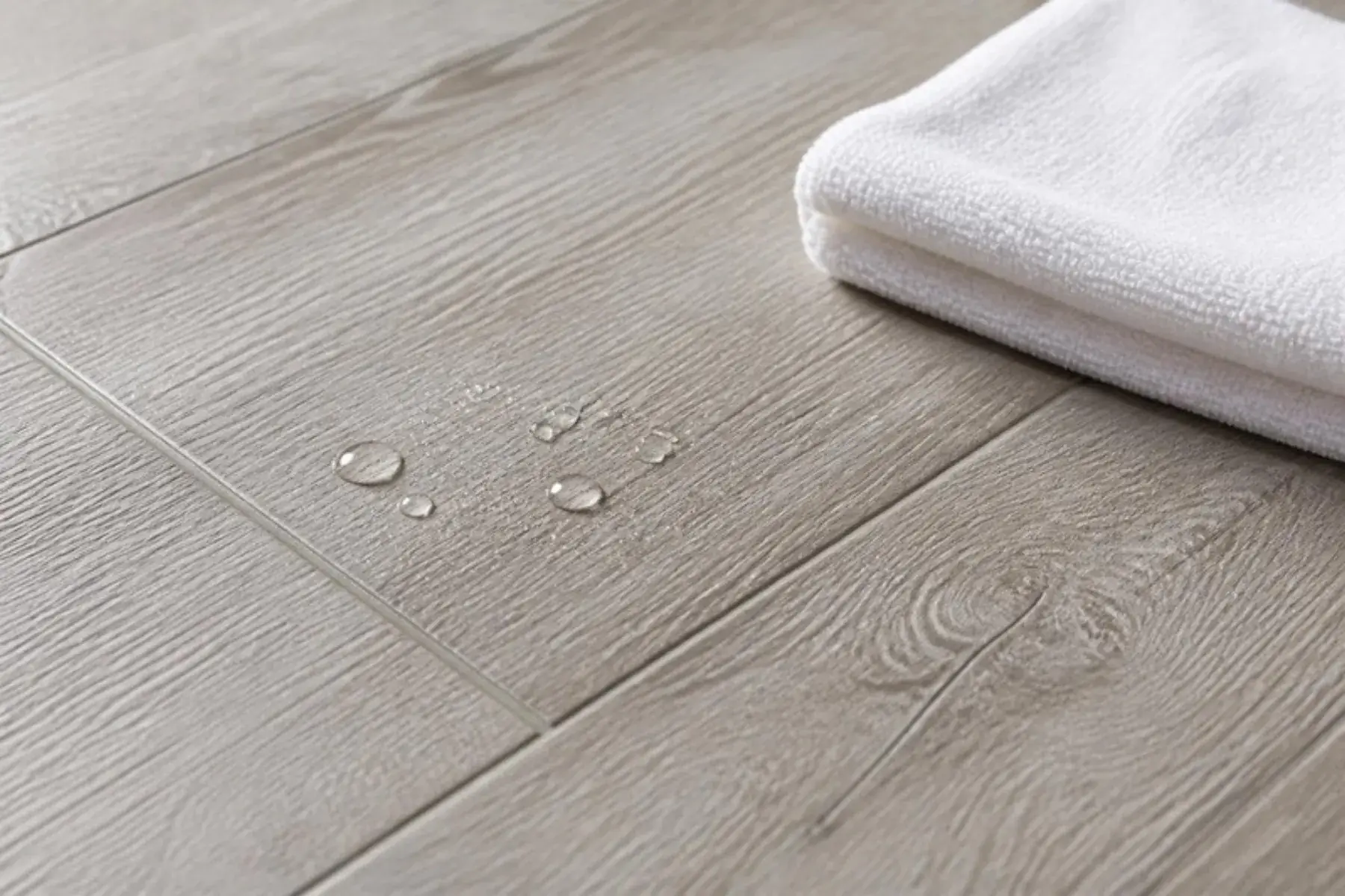 Water droplets beading on grey-washed water-resistant laminate planks with tight joints beside a microfiber cloth